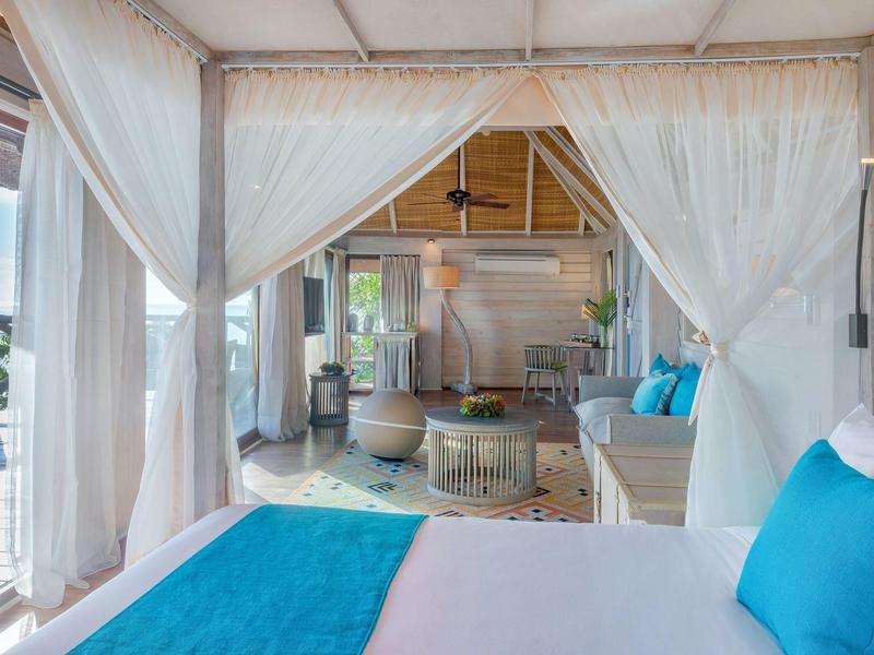 Heller, offener Raum mit Bett, blauen Kissen, gemütlicher Sitzgruppe und Vorhängen aus weißem Stoff.