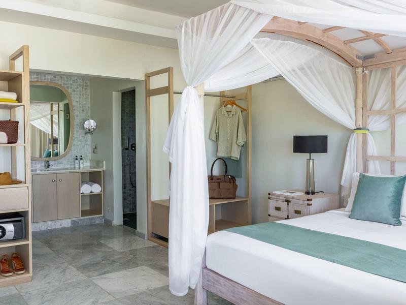 Helles Schlafzimmer mit Himmelbett, weiß-grüner Bettwäsche, kleinem Nachttisch und offenem Badezimmer.