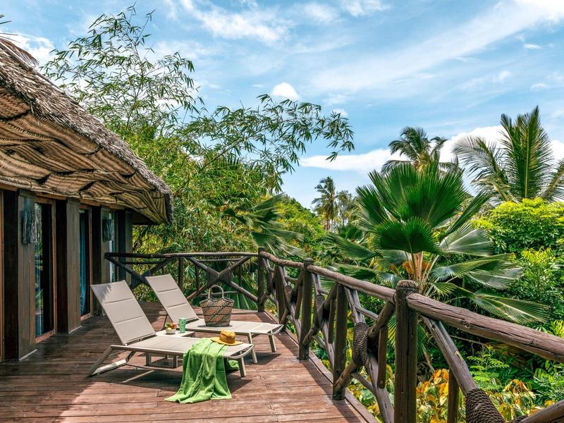 Holzterrasse mit Liegestühlen und grünem Dschungel unter blauem Himmel und Wolken.