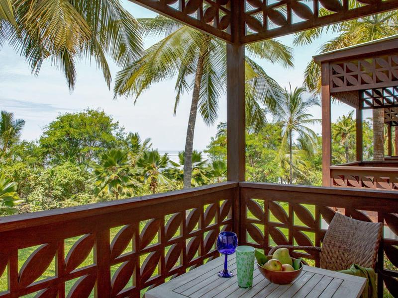 Balkon mit hölzernem Geländer, Tisch mit Flaschen und Stühlen, grüne Palmen und blauer Himmel im Hintergrund.