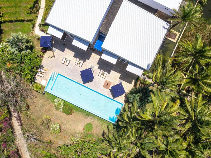 Vogelperspektive eines Hauses mit weißem Dach, Pool, Sonnenschirmen und Palmen im Garten.