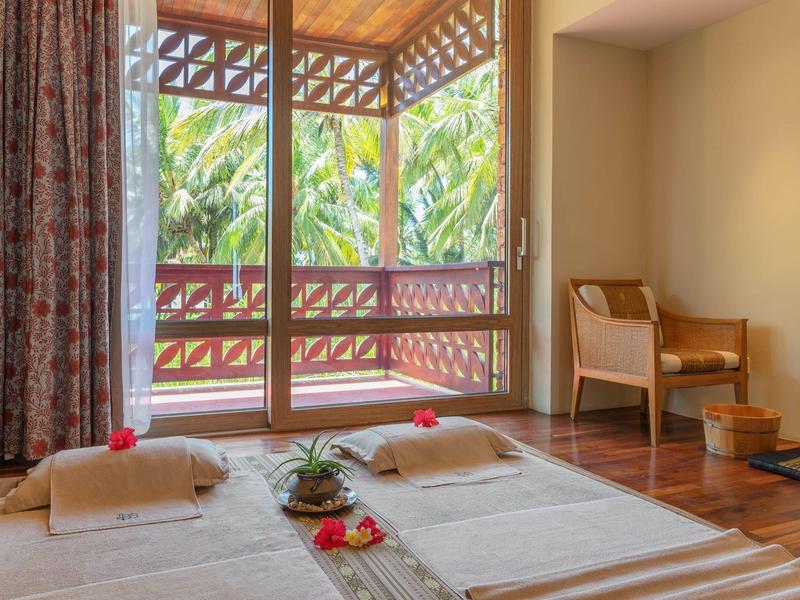 Gemütlicher Massageraum mit zwei Liegen, Holzboden, Stuhl und großer Fensterfront zum Balkon.