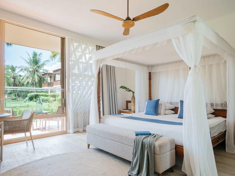 Hell eingerichtetes Schlafzimmer mit Himmelbett, Deckenventilator und Blick auf tropischen Garten.