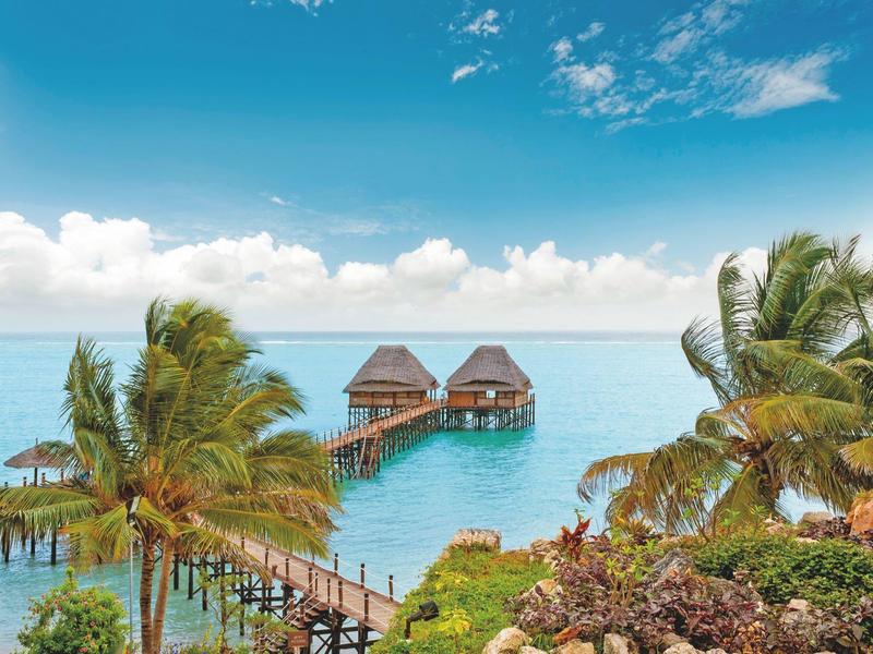 Bungalows auf Stelzen im blauen Meer mit Palmen und klarem Himmel im Hintergrund.