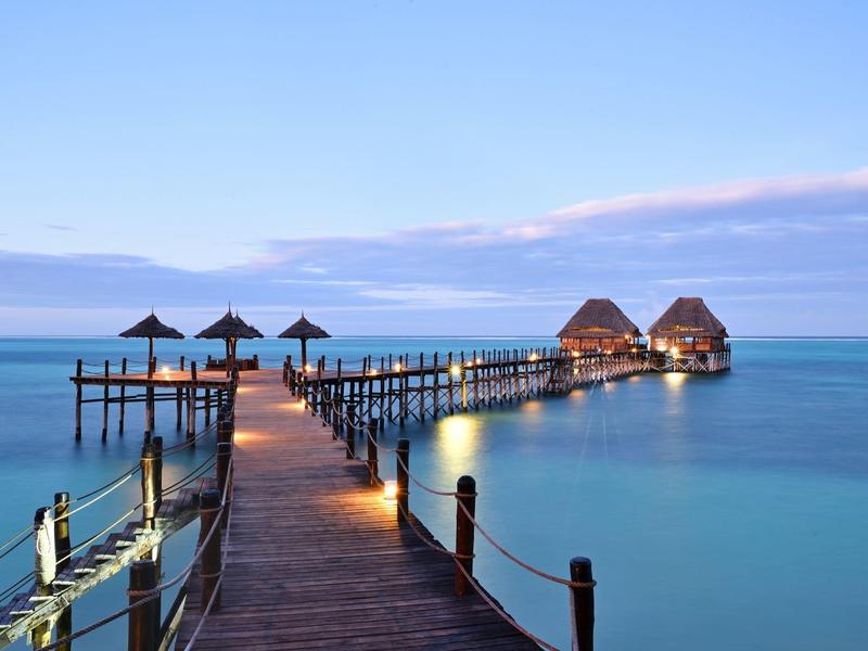 Langer Holzsteg mit Strohhütten über ruhigem, blauem Meer unter hellem Himmel bei Abenddämmerung.