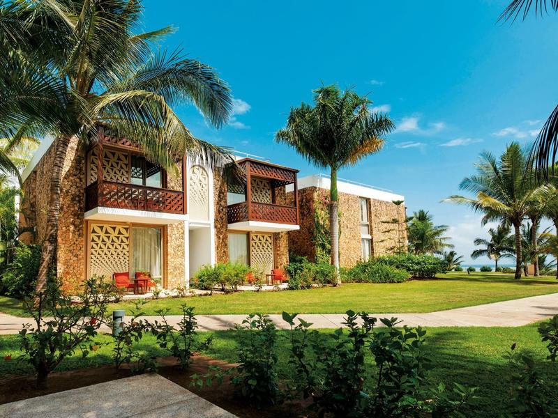 Zwei zweistöckige Häuser mit Holzbalkonen, umgeben von Palmen und grünem Rasen unter blauem Himmel.