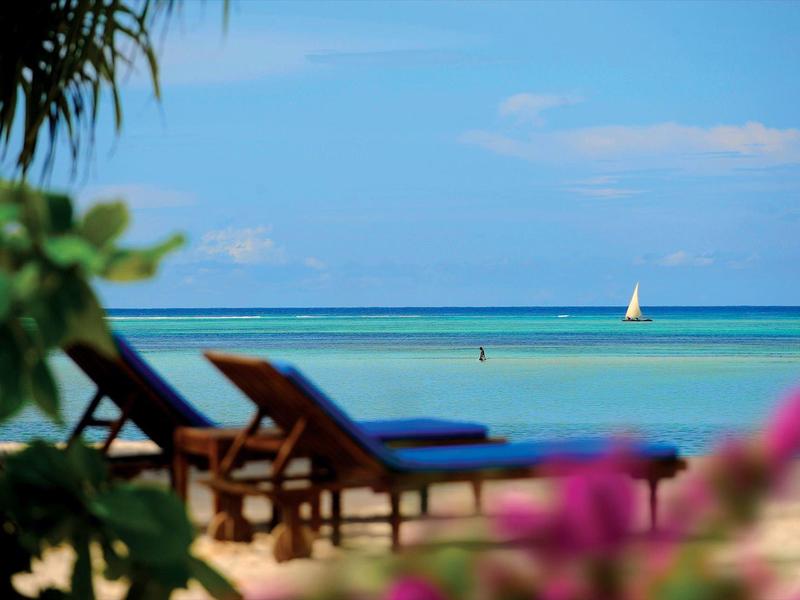 Blick auf einen Strand mit Liegestühlen, tropischen Pflanzen und Segelboot im Meer.