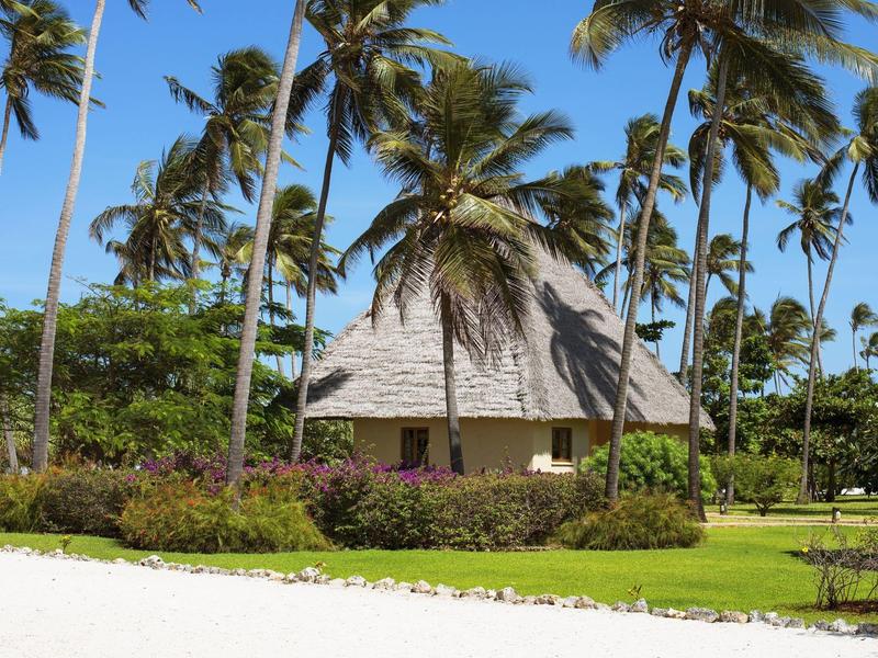 Ein Bungalow mit Strohdach umgeben von Palmen und grünem Gras am Strand.