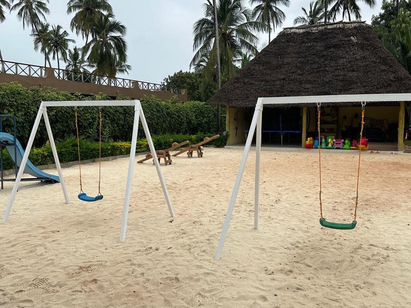 Spielplatz mit Sandboden, zwei Schaukeln und einer Rutsche vor einer Hütte und Palmen