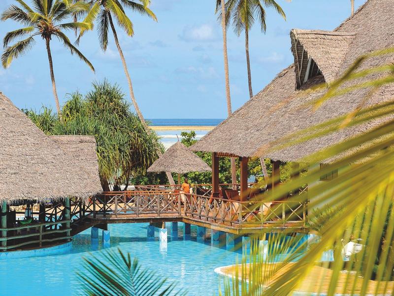 Tropisches Resort mit mehreren Poolanlagen, Palmen, Strand und grüner Landschaft bei Sonnenuntergang.