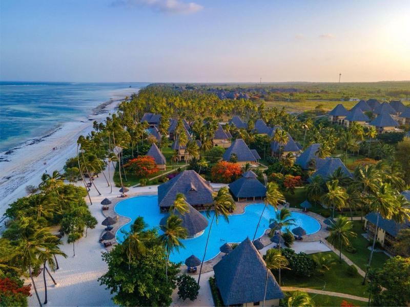 Tropisches Resort mit mehreren Poolanlagen, Palmen, Strand und grüner Landschaft bei Sonnenuntergang.