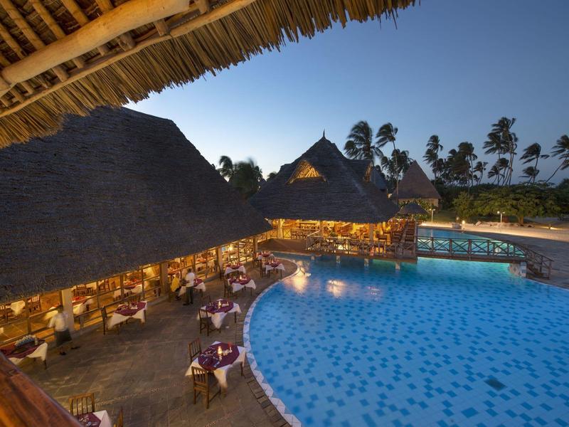 Abendaufnahme eines beleuchteten Restaurants mit Pool und Palmen unter blauem Himmel.