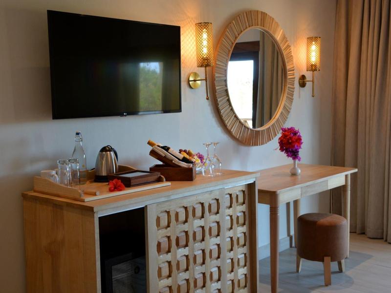 Heller Hotelzimmerbereich mit TV, Spiegel, Holzkommode und Sitzhocker neben Vorhängen.