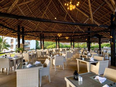 Offenes Restaurant mit Holzdach, weißen Stühlen und gedeckten Tischen in tropischer Umgebung.