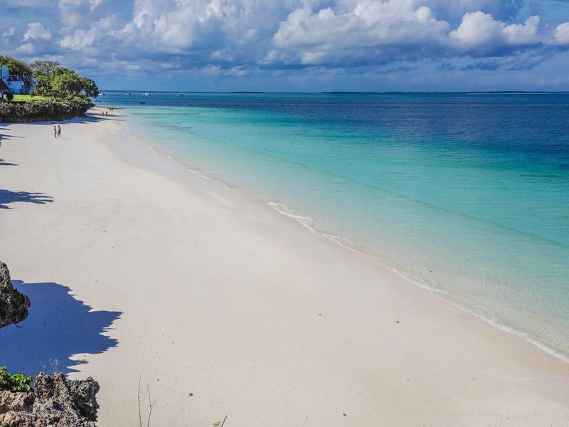 Weißer Sandstrand mit türkisem Meer und bewölktem Himmel, felsige Küste links im Bild.