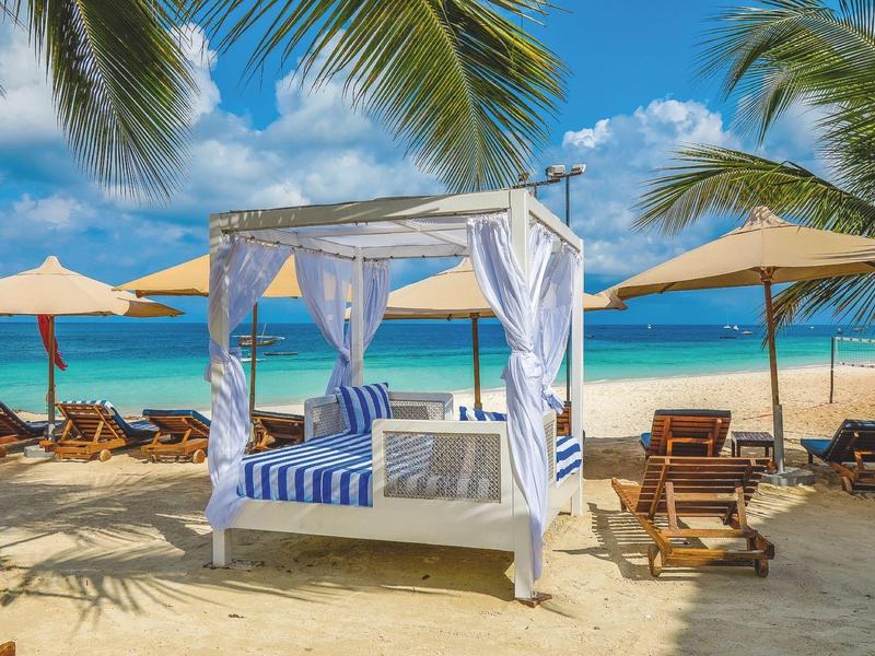 Strand mit Hängematte, Holzliegen, Sonnenschirmen, Palmen und blauem Meer im Hintergrund.