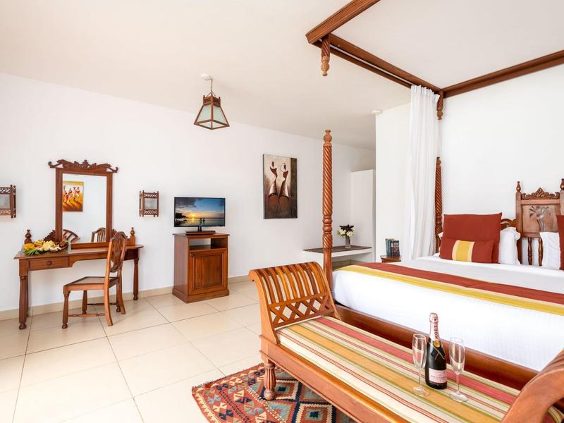 Helles Zimmer mit Holzmöbeln, großem Bett, Spiegelkommode und Bodenfliesen. Gemütlicher Teppich.