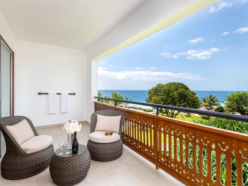 Balkon mit braunen Korbmöbeln, Holzgitter, Blick auf grüne Bäume und blauen Himmel mit Meer.