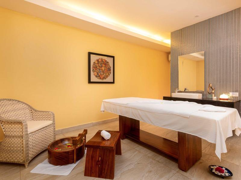 Innenraum mit Massagetisch, Korbsessel, Holzhocker und warmen gelben Wänden.