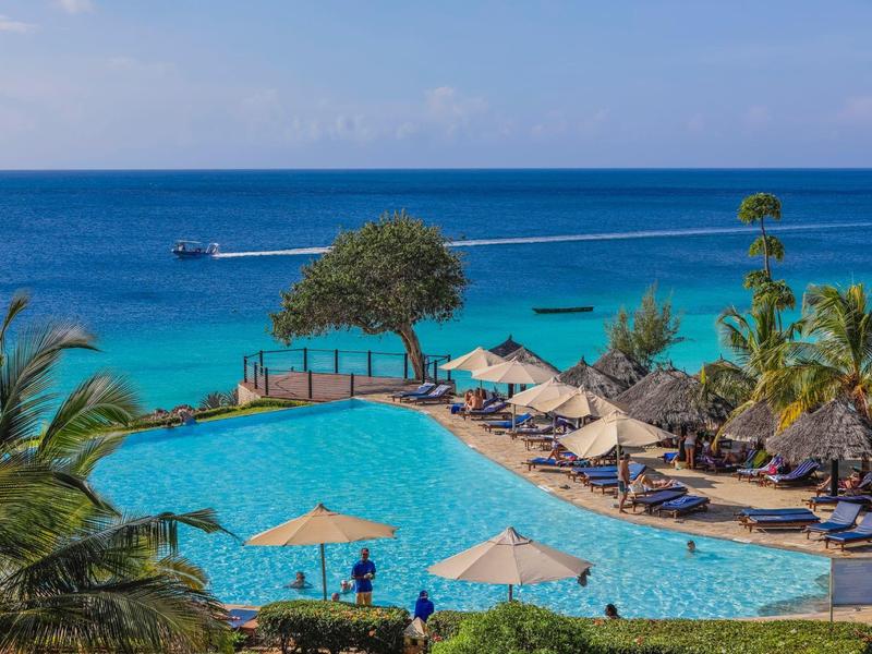 Blick auf einen großen Pool mit Sonnenschirmen, Palmen und dahinter das blaue Meer unter blauem Himmel.