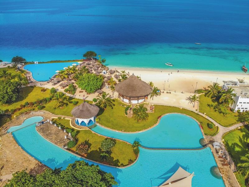 Luftaufnahme einer tropischen Küste mit türkisfarbenem Wasser, Sandstrand, Grasflächen und Wegen.