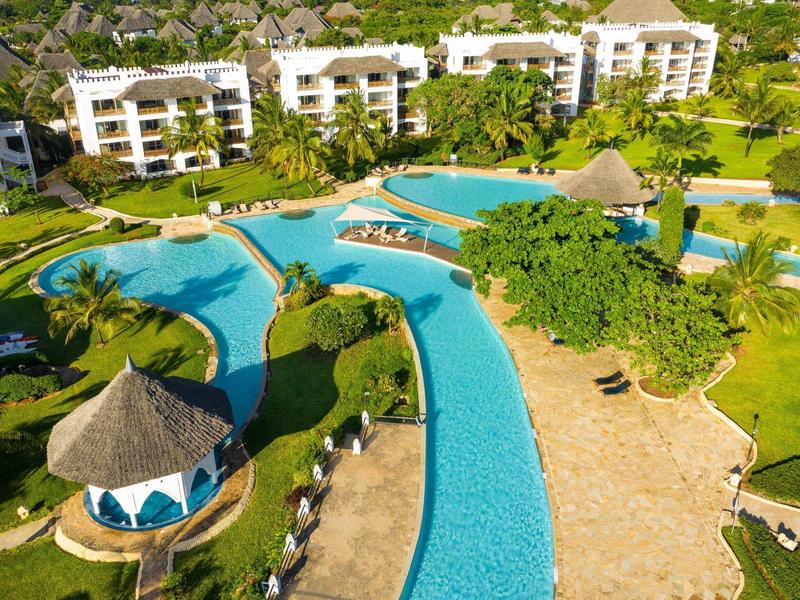 Großer, blauer Pool mit Rundwegen, Palmen und weißen Gebäuden im tropischen Resort.