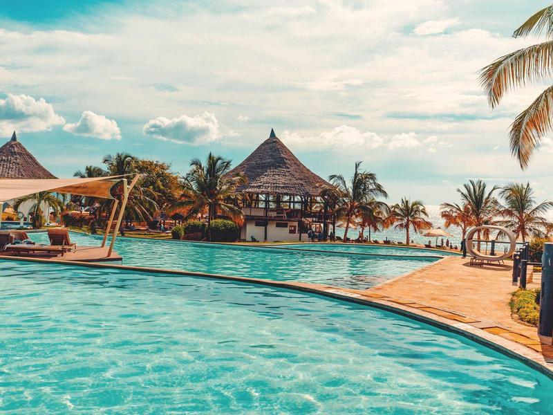 Großer Pool mit klarem Wasser, umgeben von Palmen und tropischen Pflanzen unter bewölktem Himmel.