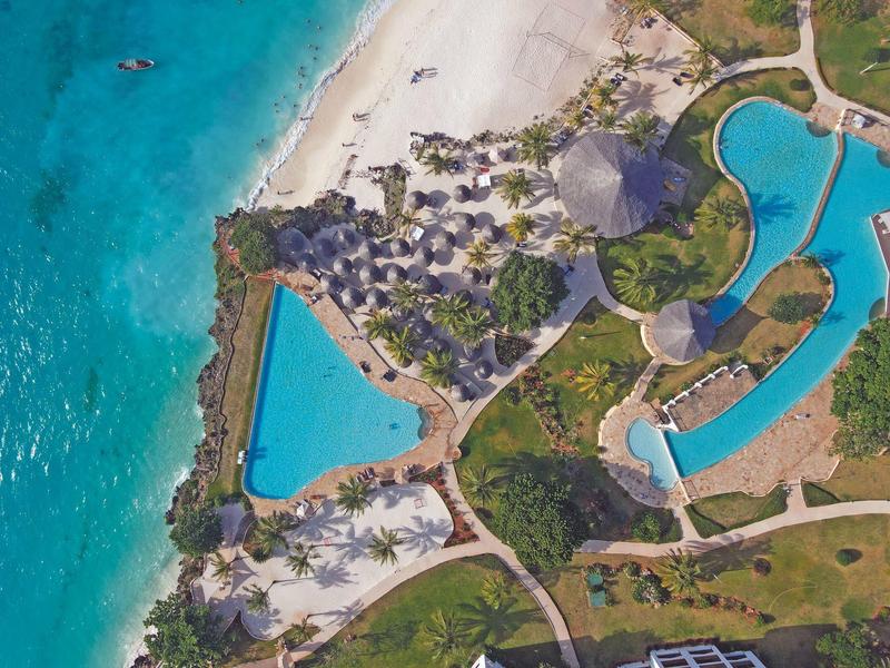 Luftaufnahme eines tropischen Resorts mit Sandstrand, blauen Pools und grünen Palmen.