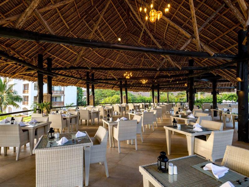 Offenes Restaurant mit Holzdach, weißen Stühlen und gedeckten Tischen in tropischer Umgebung.