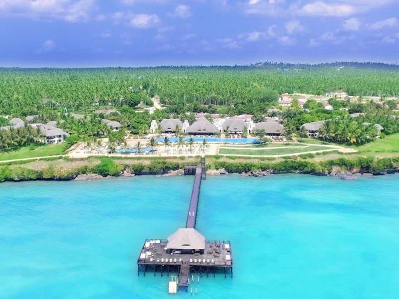 Klares blaues Meer, langer Holzsteg führt zu einer kleinen Plattform, dahinter grüner tropischer Küstenstreifen.