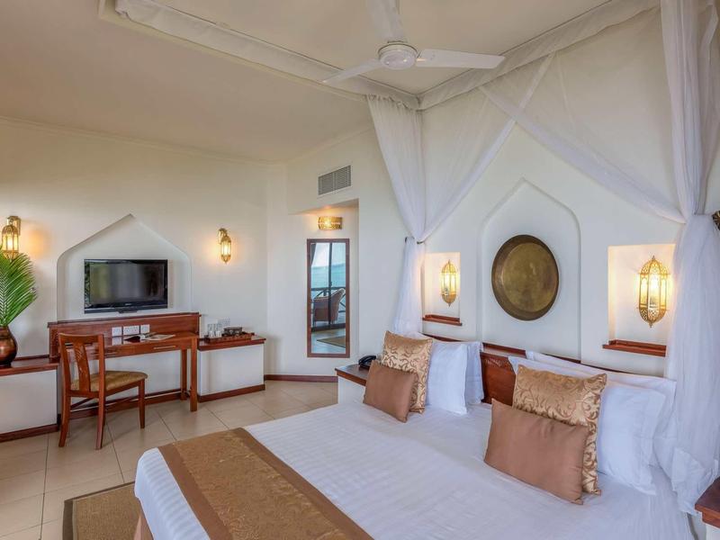 Hell eingerichtetes Schlafzimmer mit Doppelbett, Deckenventilator, Fernseher und Holzmöbeln.