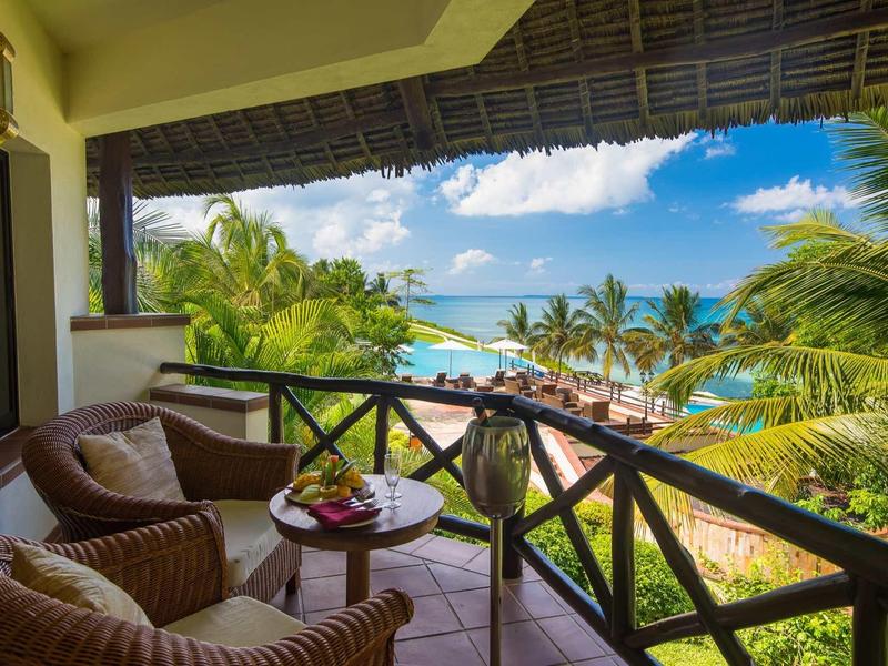 Balkon mit Korbsesseln und Tisch, Palmen, blauer Himmel und Meerblick unter bewölktem Himmel.