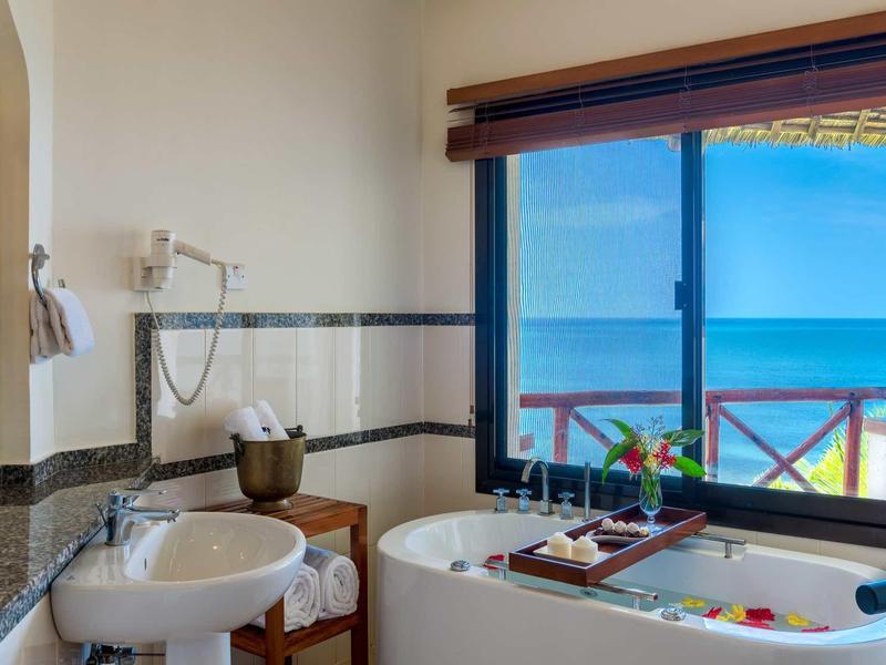 Helles Badezimmer mit Wanne, Waschbecken, Handtüchern und großem Fenster mit Meerblick.