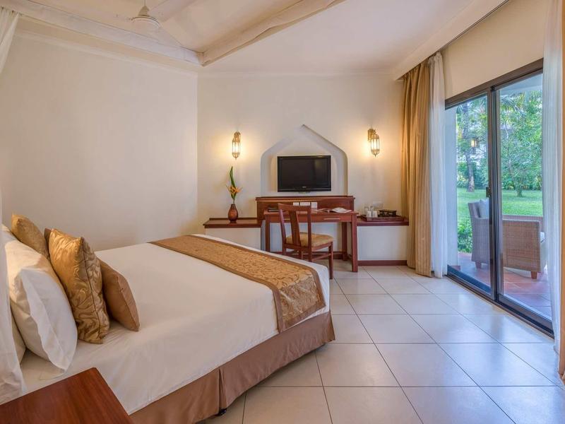 Hell eingerichtetes Hotelzimmer mit großem Bett, Schreibtisch, Fernseher und Terrassentür zum Garten.