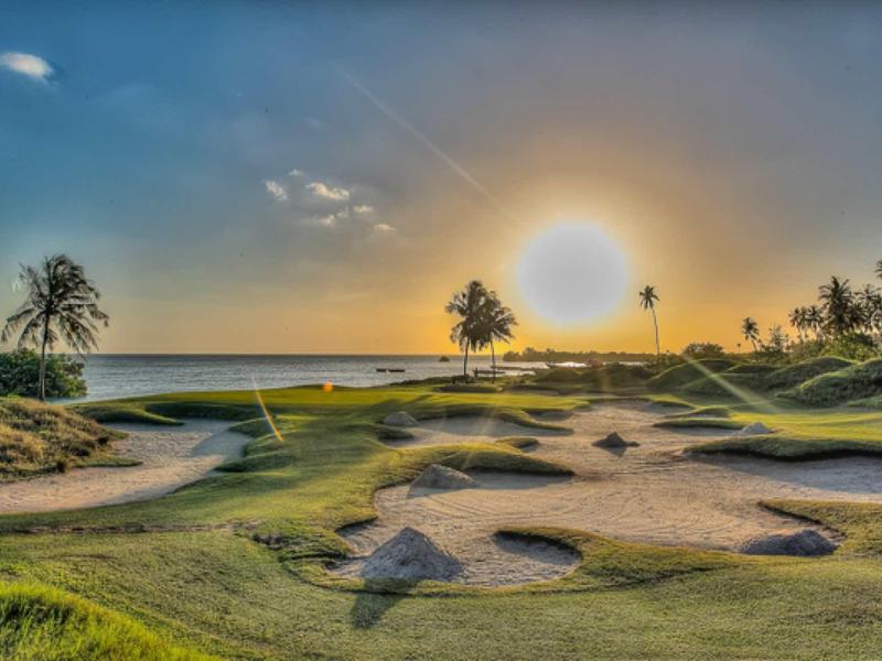 Golfplatz bei Sonnenuntergang mit Sandbunkern, grünem Gras und Palmen am Horizont.