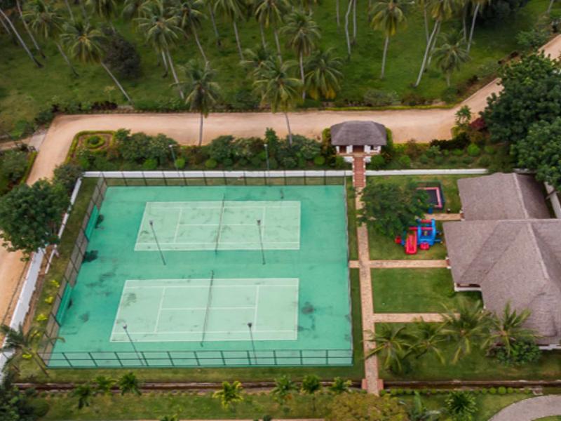 Luftaufnahme eines Tennisplatzes mit zwei Feldern, angrenzendem Haus und Kinderspielplatz in tropischer Umgebung.