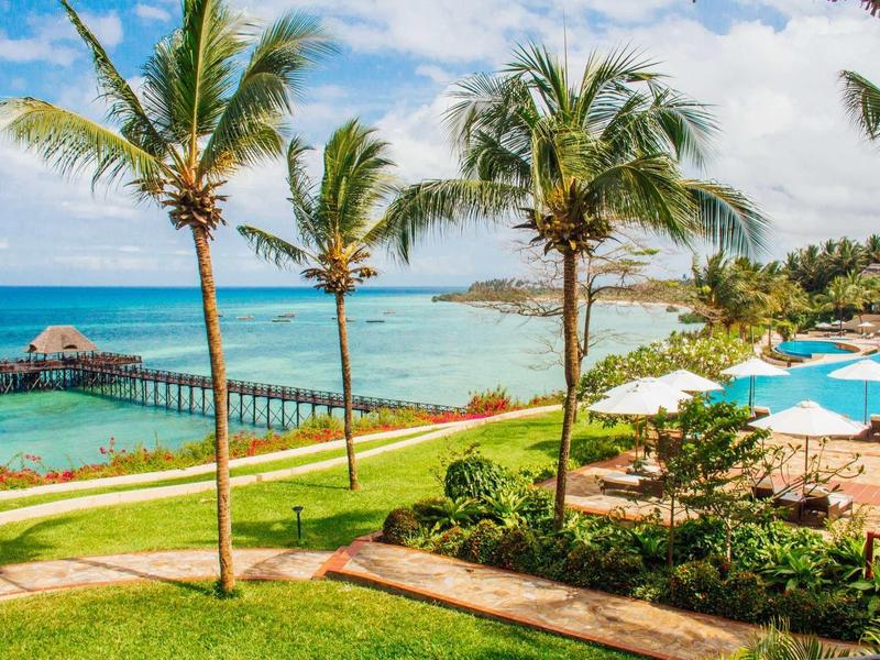 Strandresort mit Palmen, Pool, grünen Rasenflächen und Holzsteg über türkisfarbenes Meer.