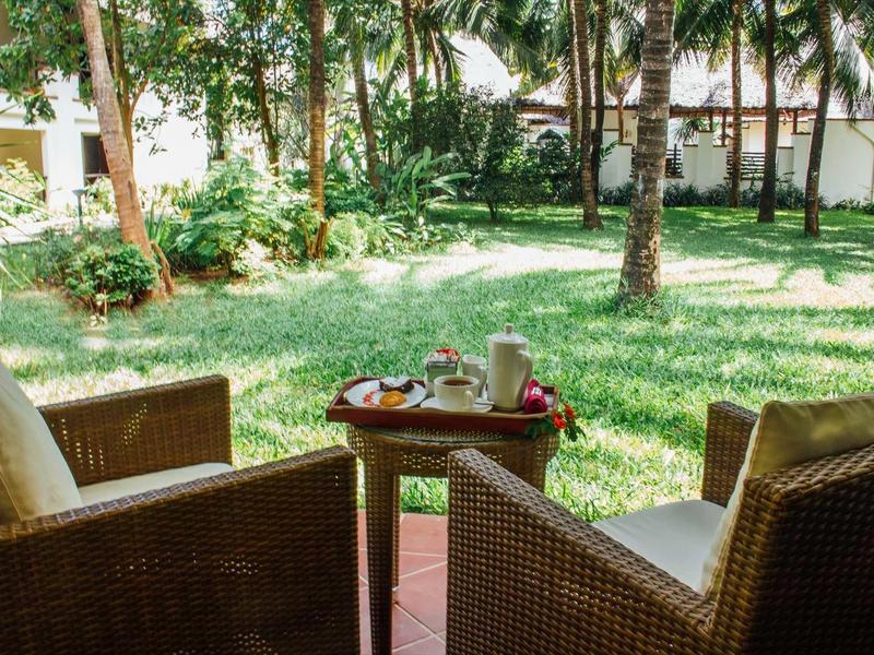 Zwei braune Korbsessel mit Polstern stehen um einen kleinen runden Tisch im Garten mit Palmen.