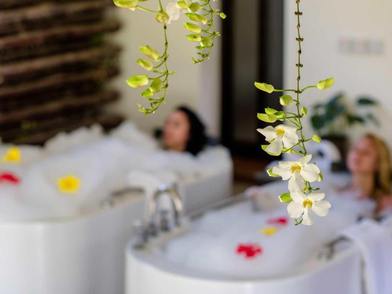 Zwei Frauen entspannen in weißen Badewannen, umgeben von Blüten und Pflanzen, Fokus auf Blüten.