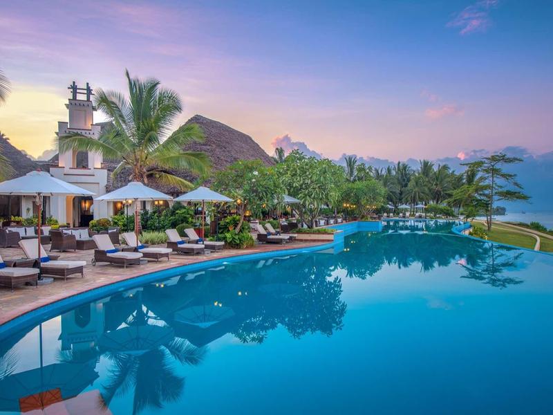 Großer Pool mit Liegestühlen, Palmen und Bergen im Hintergrund bei Sonnenuntergang.