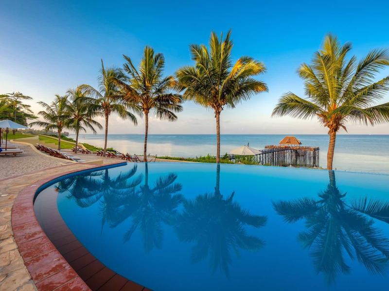 Runder Pool mit glattem Wasser und Palmen vor ruhigem Meer und blauem Himmel.