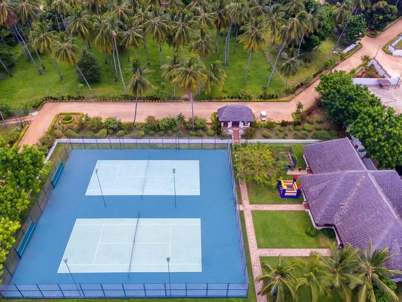 Zwei Tennisplätze mit blauen Spielfeldern, umgeben von Grün und mehreren Gebäuden auf dem Gelände.
