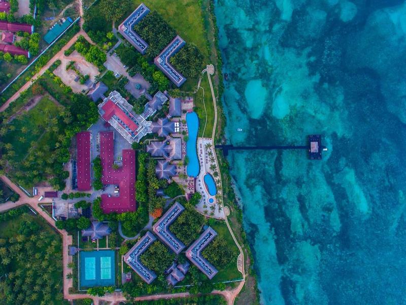 Luftaufnahme von Resort mit Gebäuden, Pool, Grünflächen und Steg ins türkisfarbene Meer.
