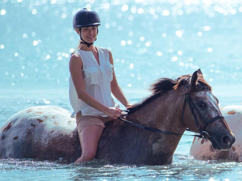 Eine Frau reitet auf einem braunen Pferd, das im blauen Wasser bis zum Bauch steht.