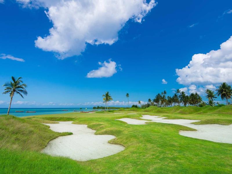 Grüner Golfplatz mit Sandbunkern, Palmen und blauem Himmel mit Wolken am Meer.