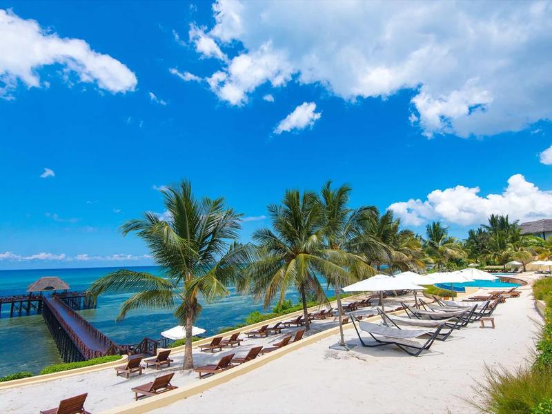 Strand mit weißem Sand, Palmen, Liegen und Sonnenschirmen unter blauem Himmel und Wolken.