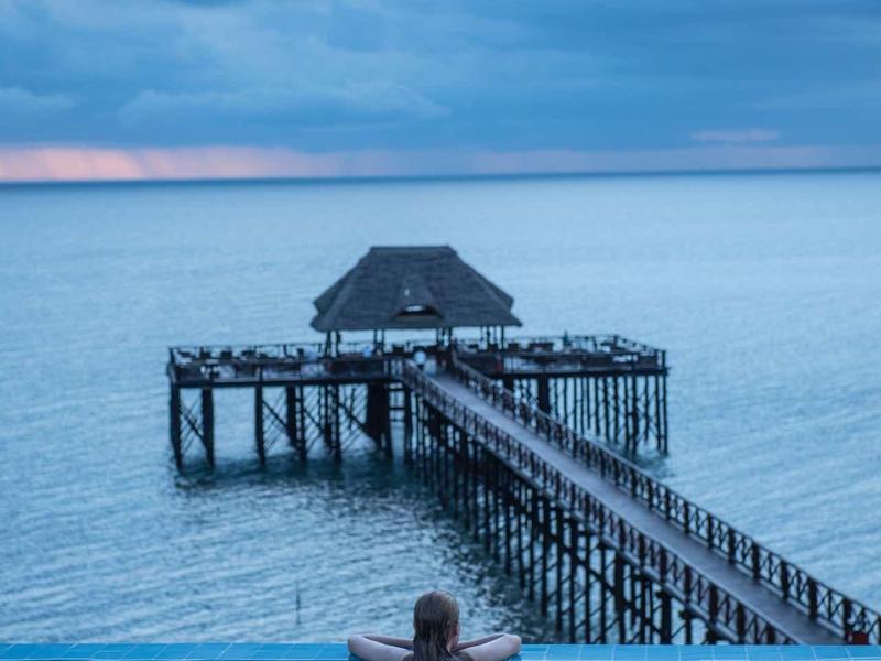 Person schwimmt im Infinity-Pool mit Blick auf Steg und ruhiges Meer unter bewölktem Himmel.