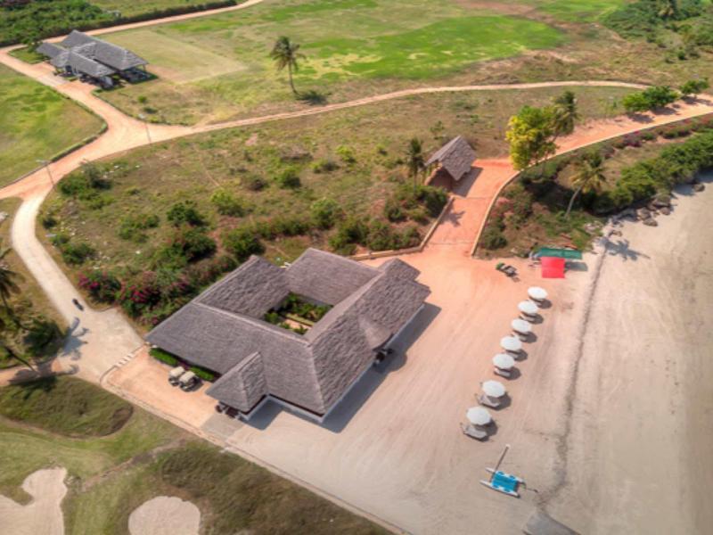 Luftaufnahme eines Strandhauses mit Roofdach, Sonnenschirmen und Sandstrand am grünen Gelände.