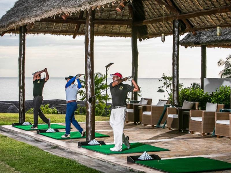 Drei Personen schlagen Golfbälle unter einem strohgedeckten Pavillon mit Blick aufs Wasser.