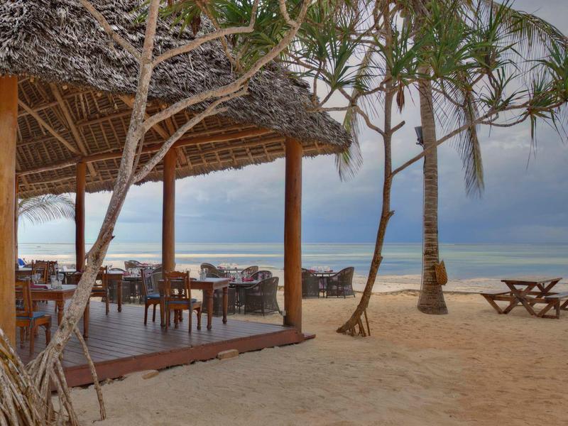 Offener Strandpavillon mit Essbereich und Sitzgelegenheiten auf sandigem Boden neben dem Meer.